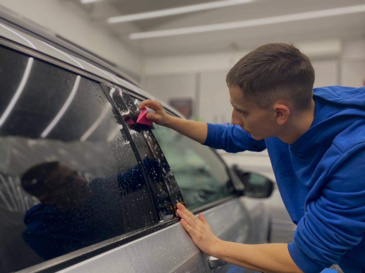 Pracownik w niebieskiej bluzie aplikuje folię na szybę srebrnego auta, używając różowego aplikatora. Widoczne krople wody.