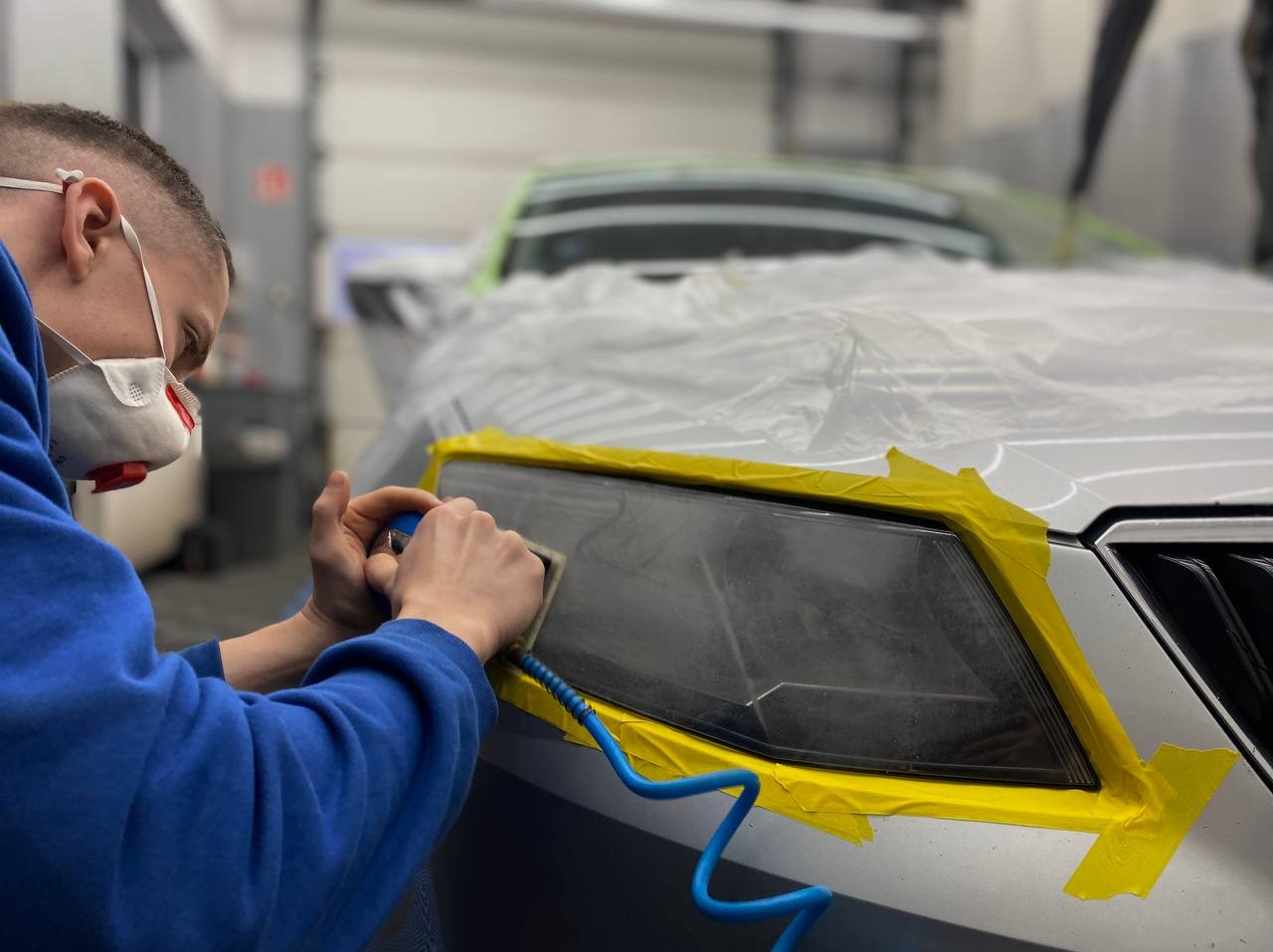 Pracownik w Łodzi poleruje reflektor samochodu zabezpieczony taśmą, używając szlifierki pneumatycznej. Auto owinięte folią ochronną.