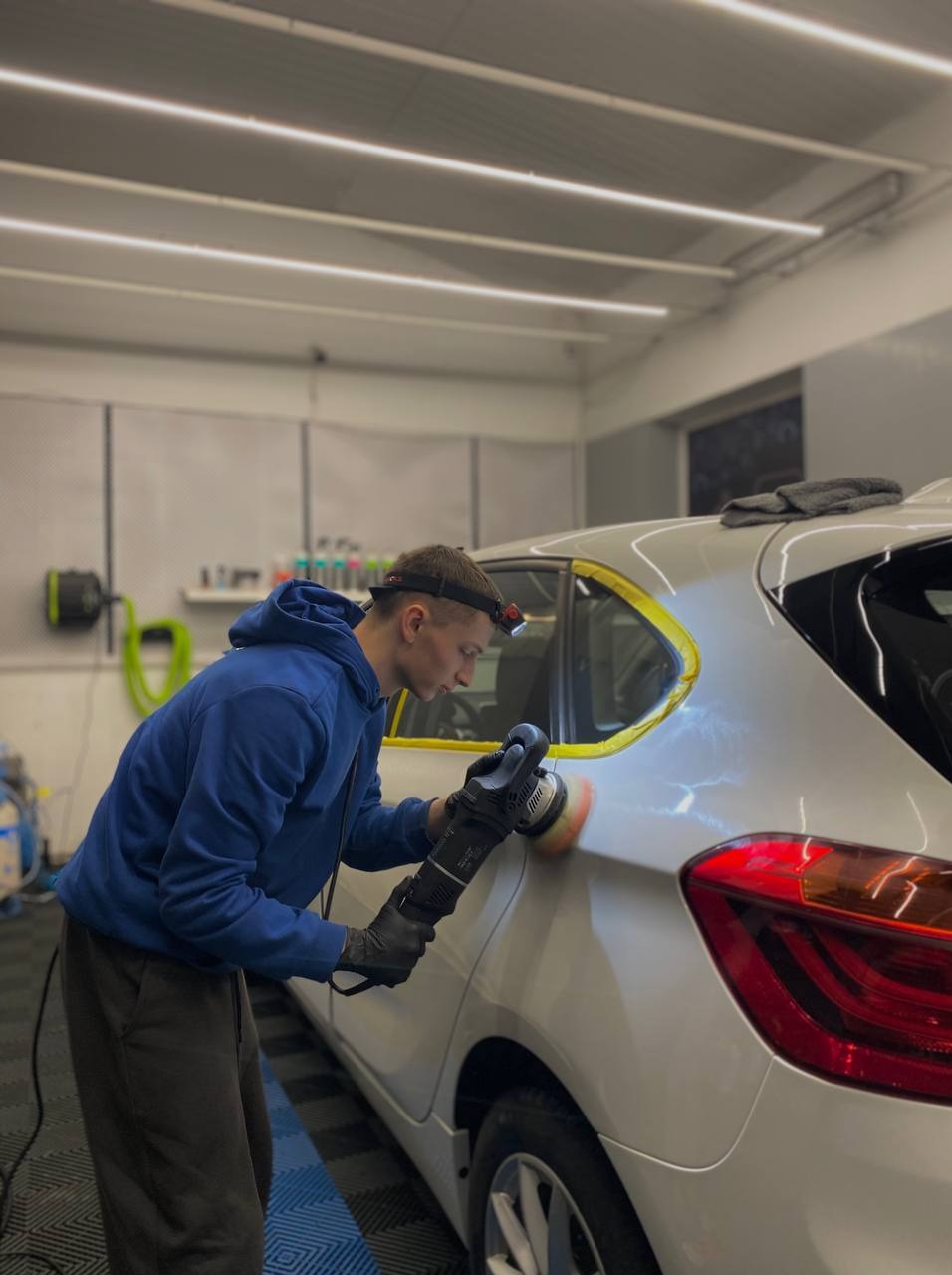 Detailing auta: mężczyzna z lampką na głowie poleruje biały samochód z żółtą taśmą ochronną, używając polerki mechanicznej w dobrze oświetlonym garażu.