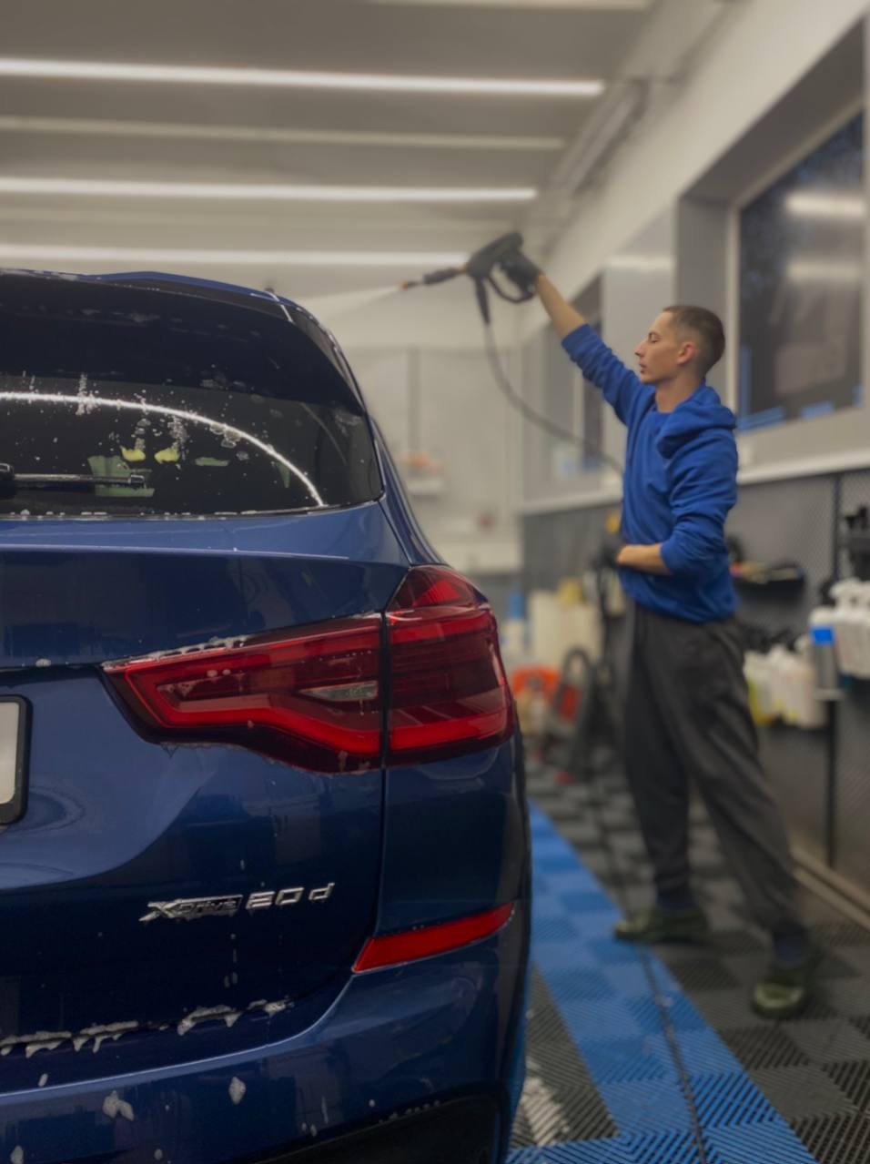 Mycie ciśnieniowe niebieskiego BMW X3 w myjni. Widok z tyłu, w tle osoba z lancą. Błyszcząca karoseria, profesjonalne oświetlenie, czysta posadzka. Dbałość o detale.