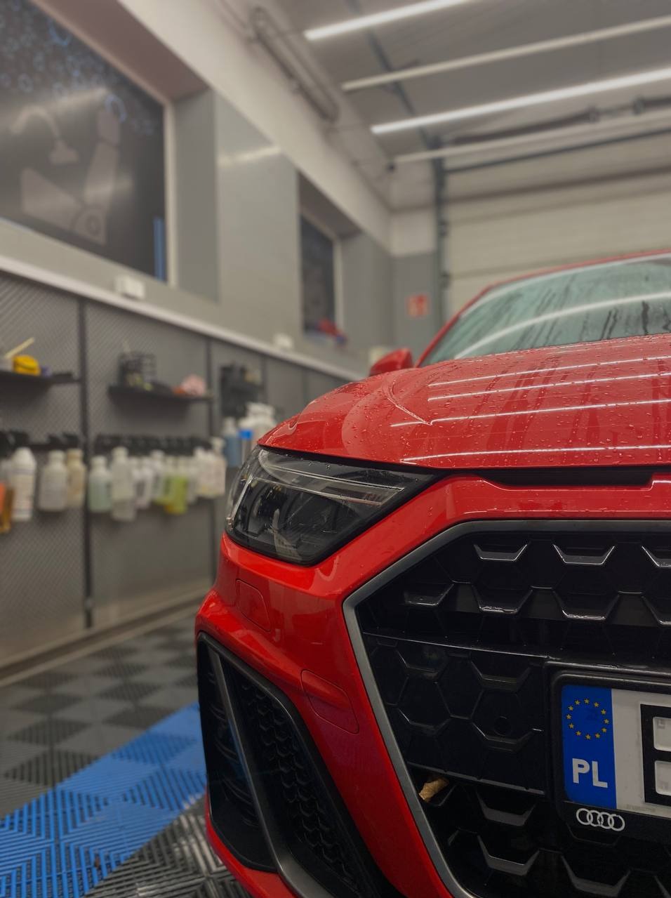 Czerwone Audi A1 po myciu, krople wody na karoserii, widok na reflektor i grill, tablica rejestracyjna PL, wnętrze myjni z półkami i akcesoriami w tle.