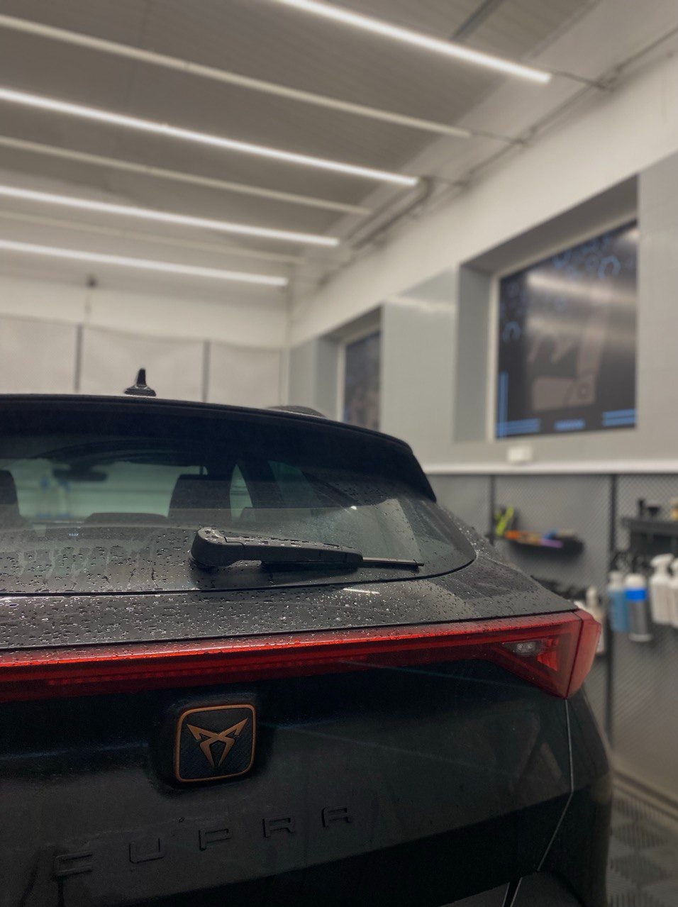 Czarny samochód Cupra po myciu, z widocznymi kroplami wody na karoserii, w jasnym pomieszczeniu myjni. Widoczne logo marki i tylna wycieraczka.