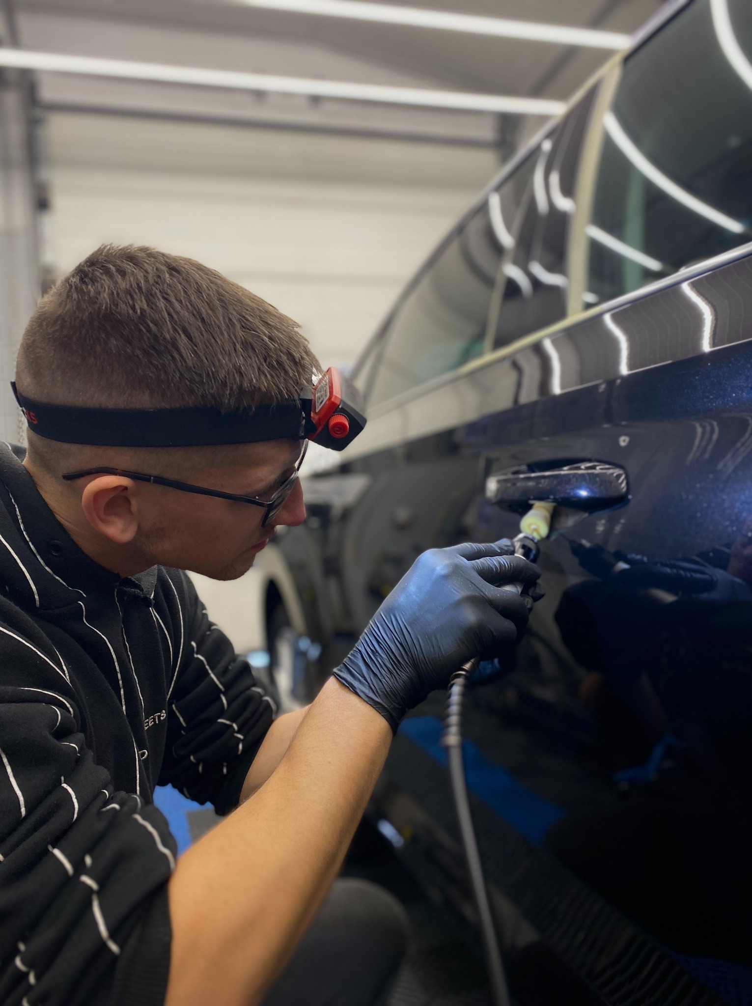 Detailing auta: polerowanie lakieru wokół klamki ciemnego samochodu przez detailera w rękawicach, z lampą na głowie, w warsztacie.