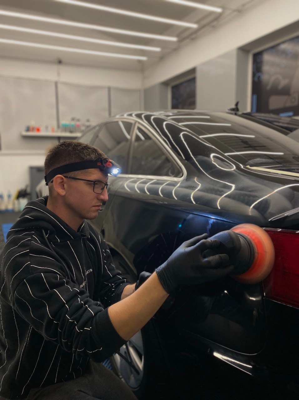 Polerowanie tylnej lampy czarnego auta przez mężczyznę w czarnej bluzie i okularach, z lampą czołową, w dobrze oświetlonym garażu.