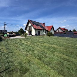 MANDRAGORA KAROL JAGNICKI - Rozległy trawnik z rolki przed domem z panelami słonecznymi, widziany w słoneczny dzień. Widoczny sprzęt ogrodniczy i osoby pracujące w tle.