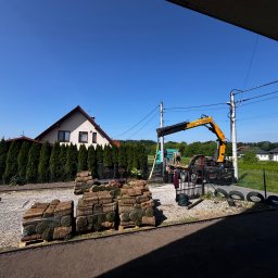 MANDRAGORA KAROL JAGNICKI - Układanie trawy z rolki: palety z trawą, żuraw, przygotowany teren. Dom z czerwonym dachem w tle, słoneczny dzień.