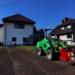 MANDRAGORA KAROL JAGNICKI - Przygotowanie gruntu pod trawnik: zielony miniciągnik Avant z frezem glebowym na uprawionej ziemi, dom w tle w słoneczny dzień.