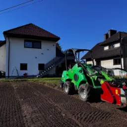Przygotowanie gruntu pod trawnik: zielony miniciągnik Avant z frezem glebowym na uprawionej ziemi, dom w tle w słoneczny dzień.