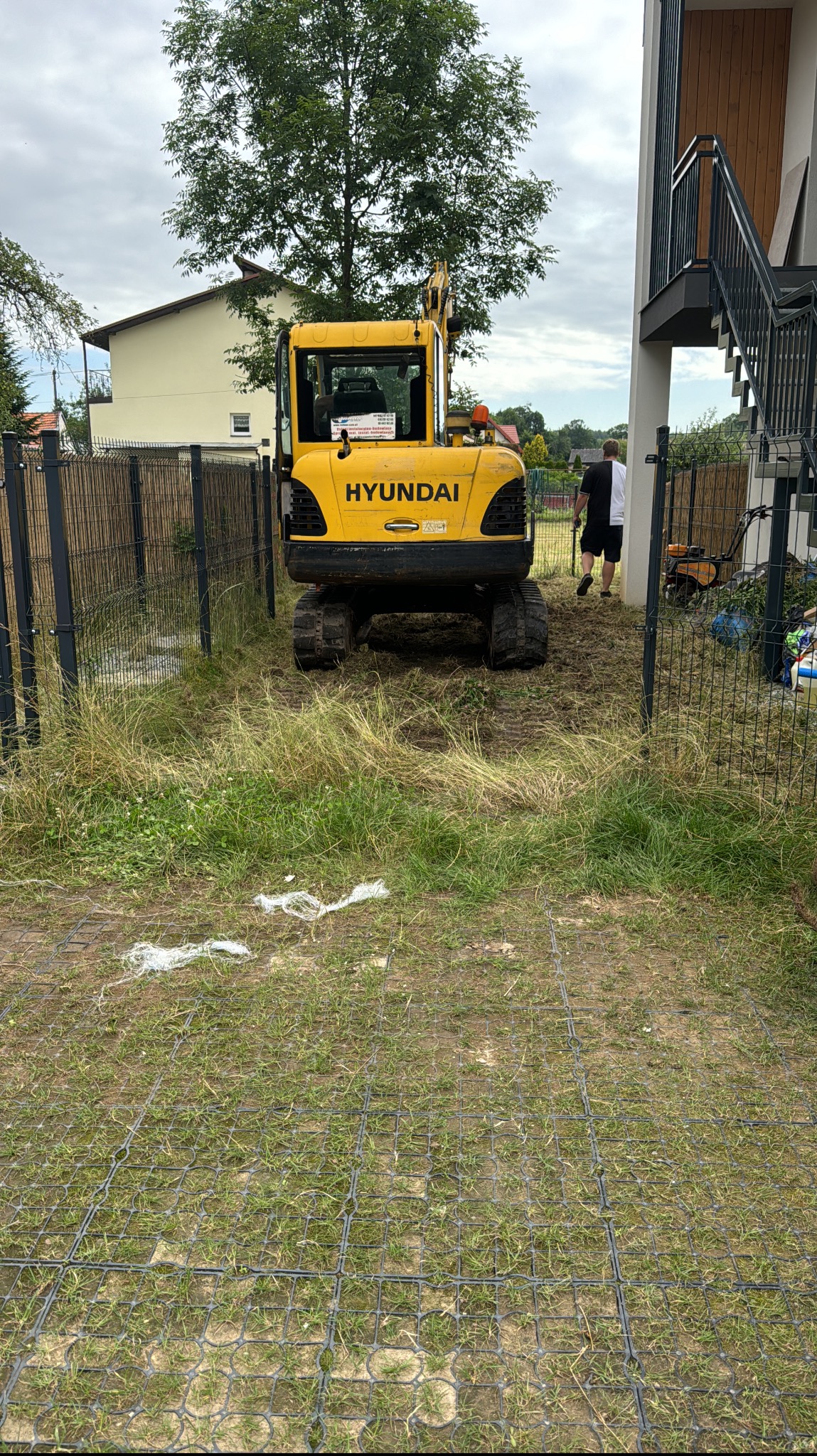 Żółta koparka Hyundai na trawniku z plastikową kratką, przygotowująca teren pod trawnik z rolki, widoczny fragment domu i ogrodzenie.