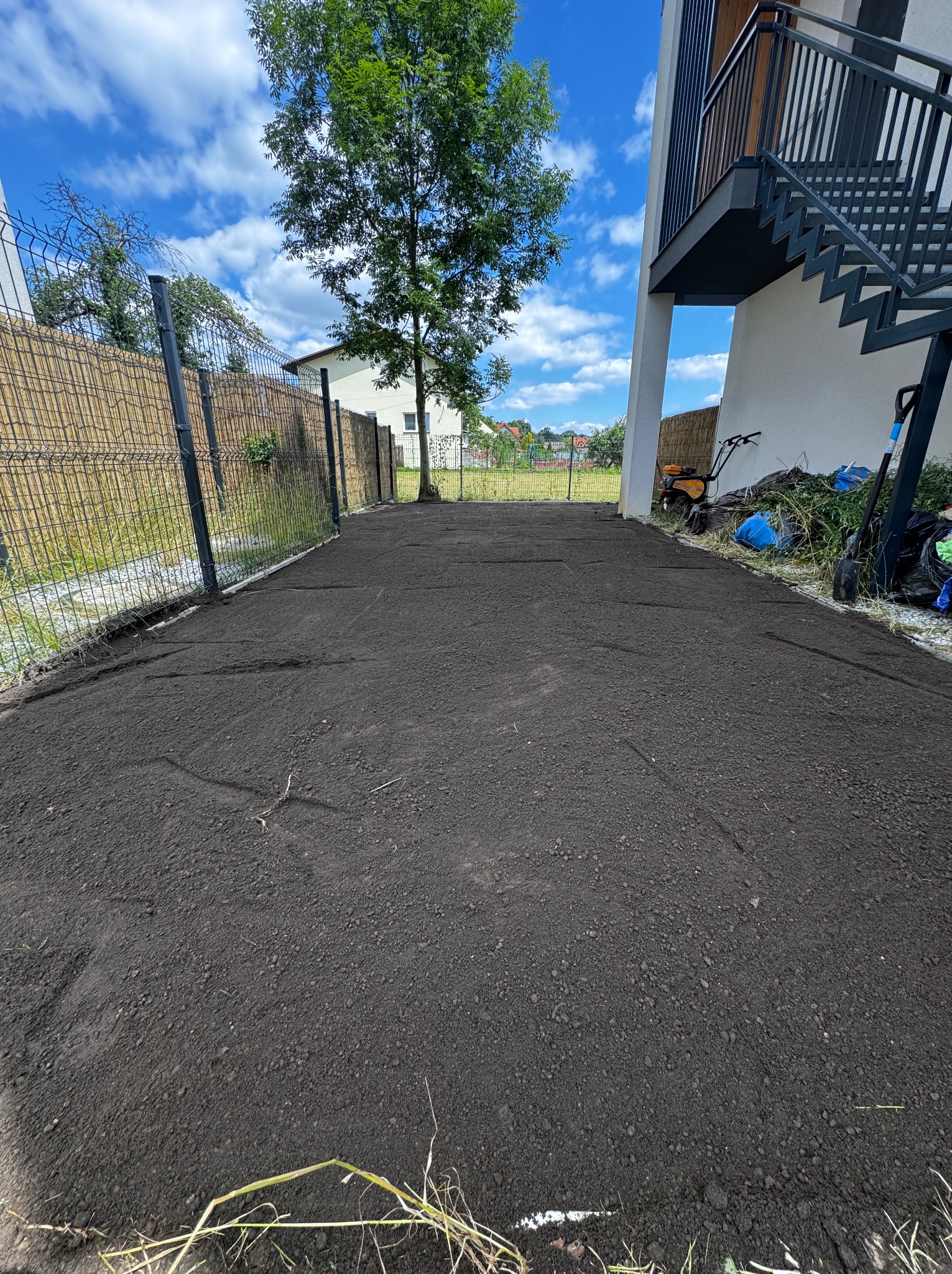 Przygotowany teren pod trawnik: wyrównana, ciemna ziemia czeka na rozłożenie trawy z rolki, widoczne ślady po wałowaniu, w tle ogrodzenie z siatki i budynek z zewnętrznymi schodami.