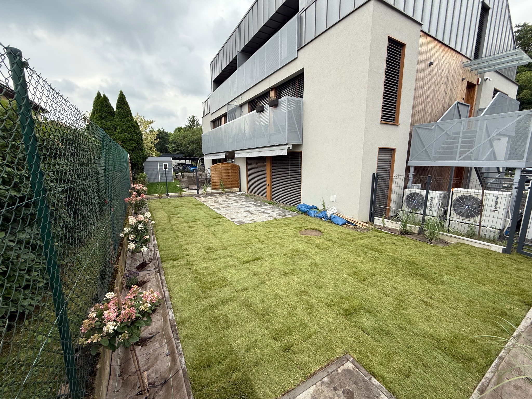 Świeżo ułożona, soczysta zieleń trawy z rolki na tle nowoczesnego budynku z balkonami i ogrodzeniem z kwiatami. Widoczne klimatyzatory i fragment kostki brukowej.