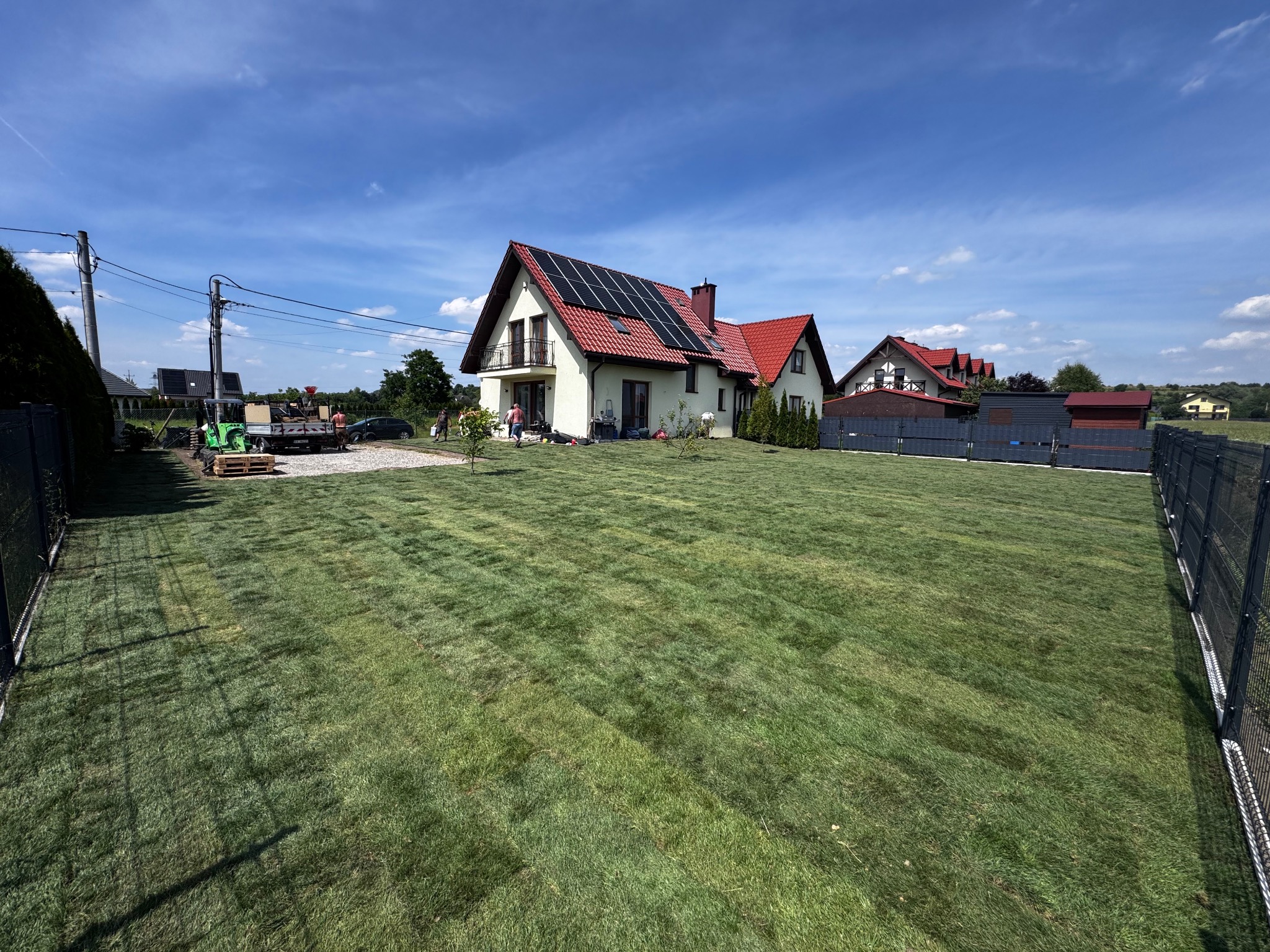 Rozległy trawnik z rolki przed domem z panelami słonecznymi, widziany w słoneczny dzień. Widoczny sprzęt ogrodniczy i osoby pracujące w tle.