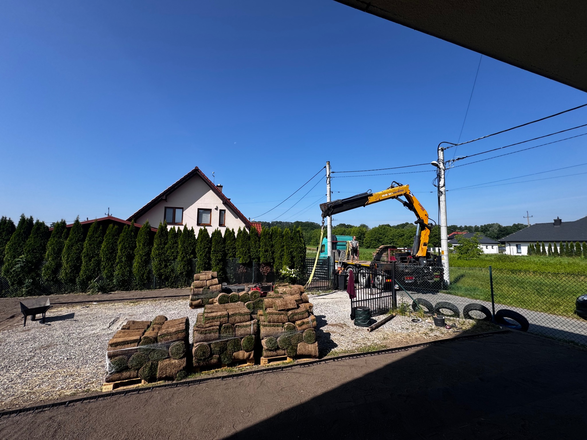 Układanie trawy z rolki: palety z trawą, żuraw, przygotowany teren. Dom z czerwonym dachem w tle, słoneczny dzień.