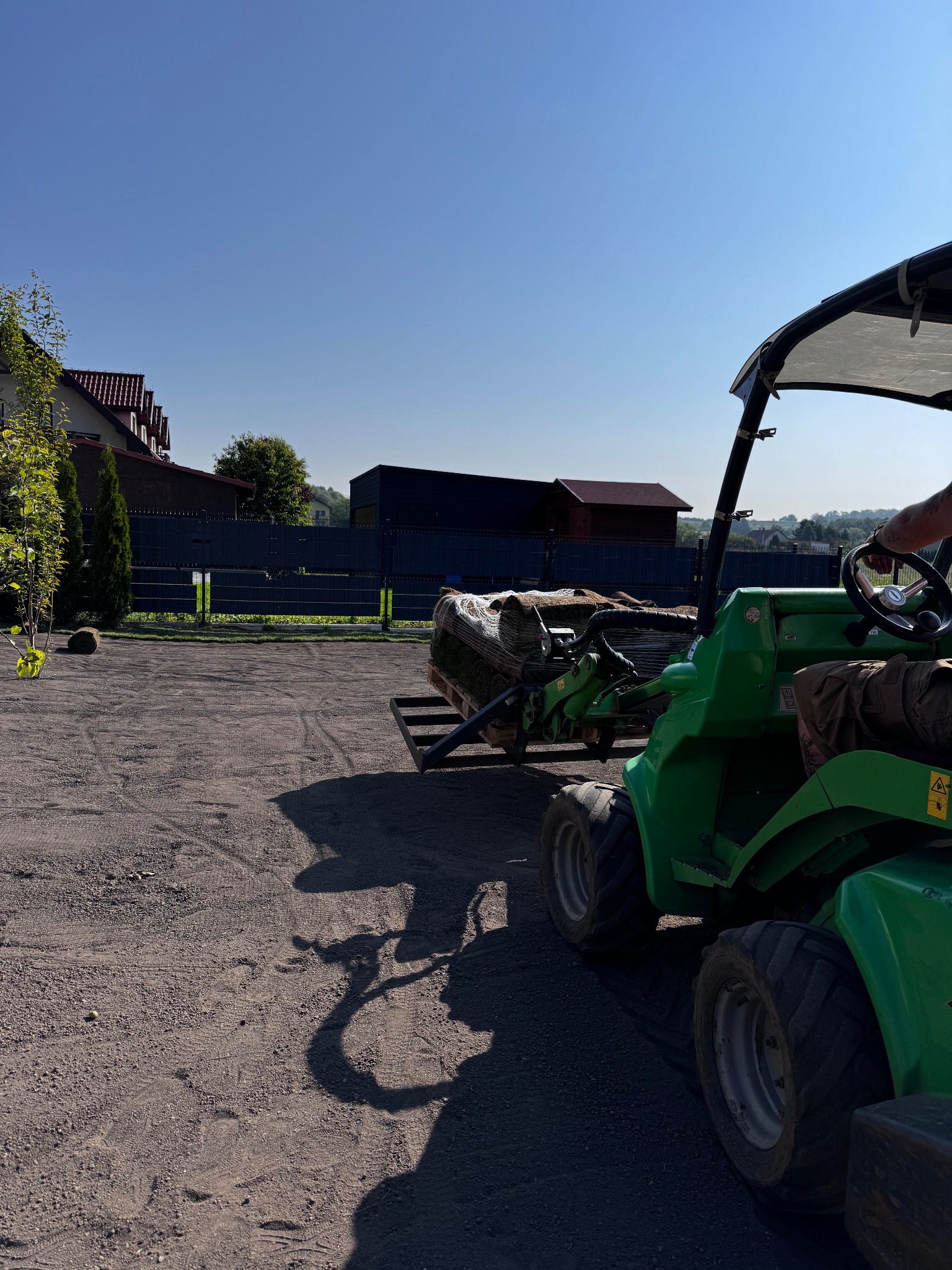 Zielony miniładowarka Avant transportuje paletę z trawą z rolki na przygotowany teren podwórza, w tle domy i ogrodzenie. Słoneczny dzień.