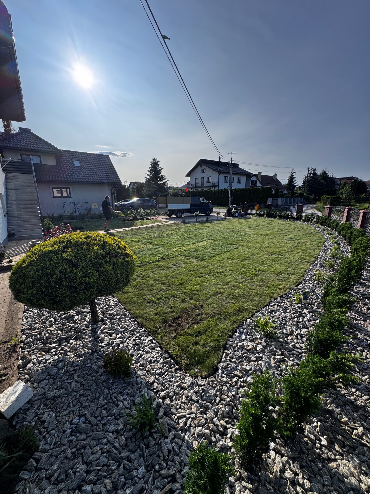 Ułożony trawnik z rolki, obsypany kamieniami ozdobnymi, z niskimi krzewami wzdłuż obrzeży. W tle domy i osoba pracująca w ogrodzie.