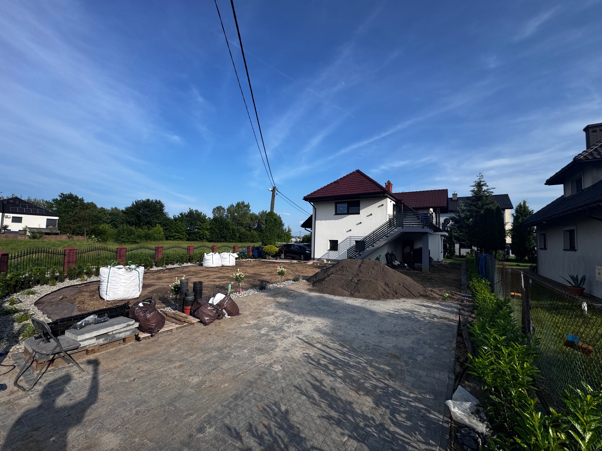 Przygotowanie terenu pod ogród: worki z ziemią, kamienie, obrzeża, usypana ziemia na posesji z kostką brukową. Widok na dom i zieleń w tle.