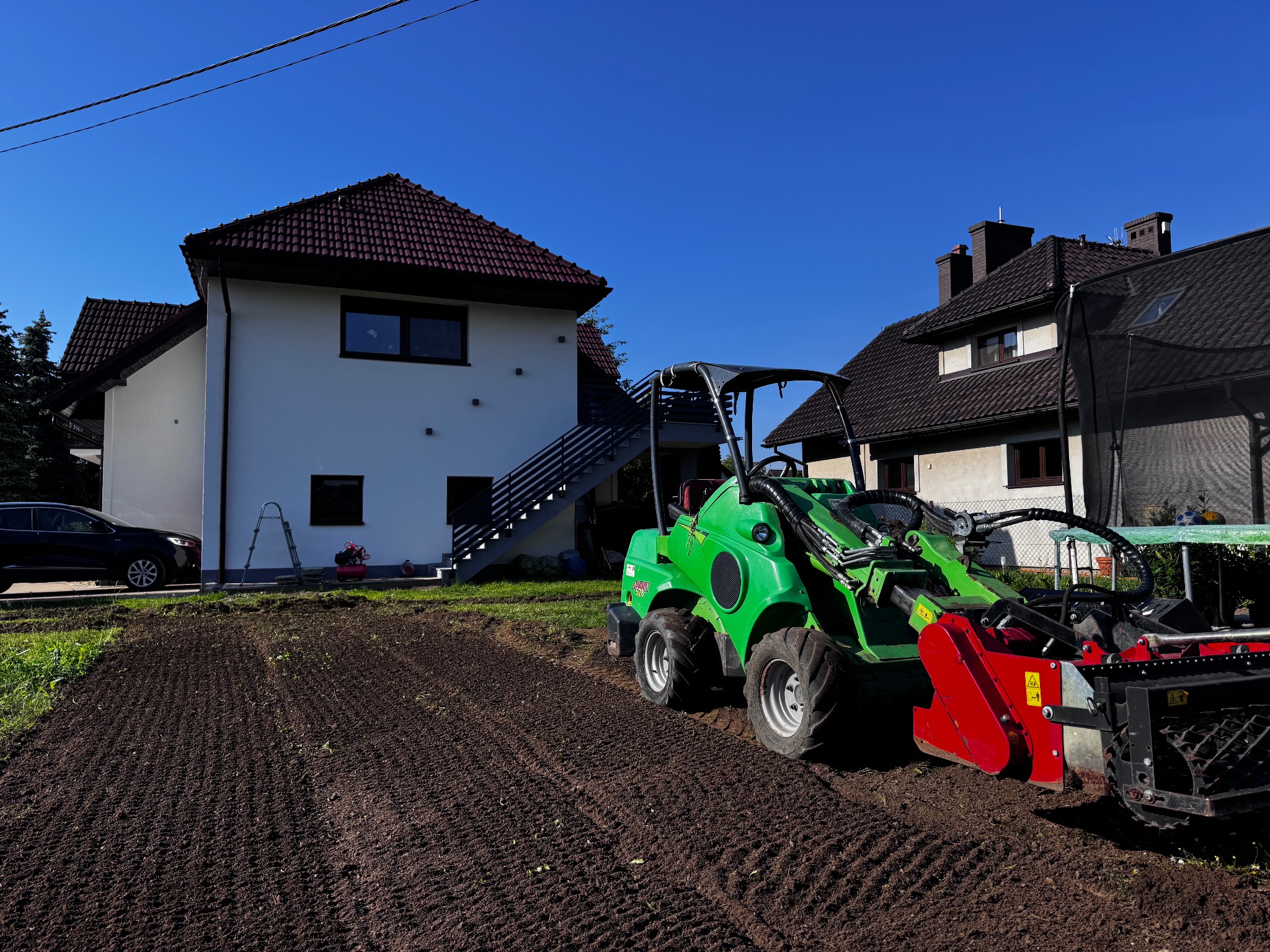 Przygotowanie gruntu pod trawnik: zielony miniciągnik Avant z frezem glebowym na uprawionej ziemi, dom w tle w słoneczny dzień.