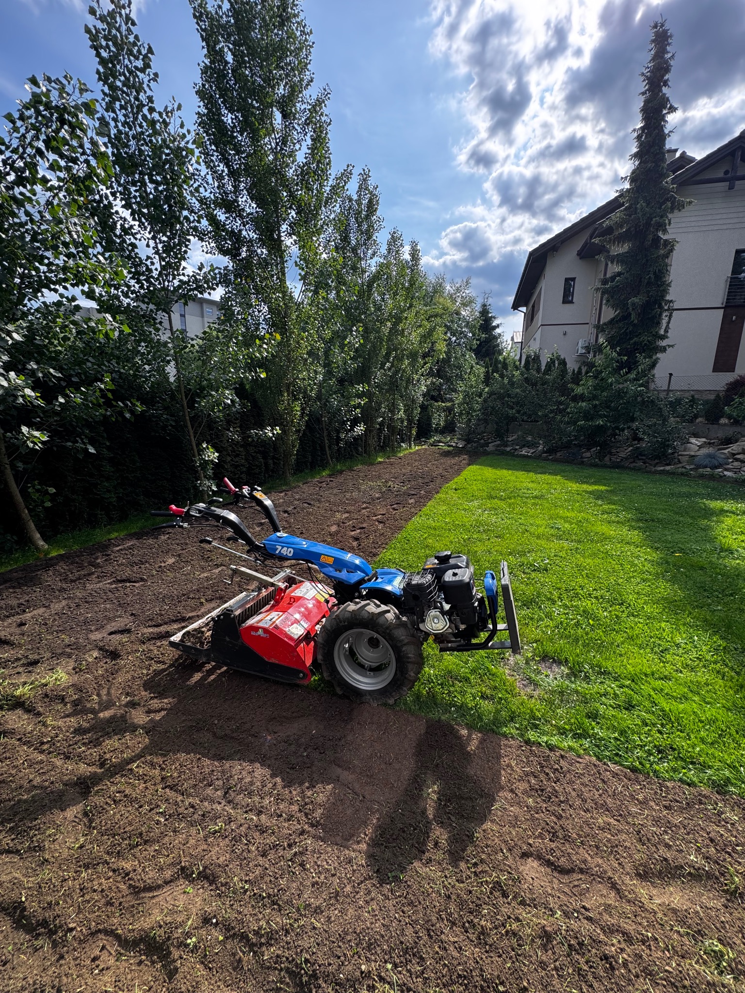 Niebiesko-czerwony glebogryz na przygotowanym podłożu, obok zielony trawnik. Widoczne drzewa i budynek w tle. Słoneczny dzień.