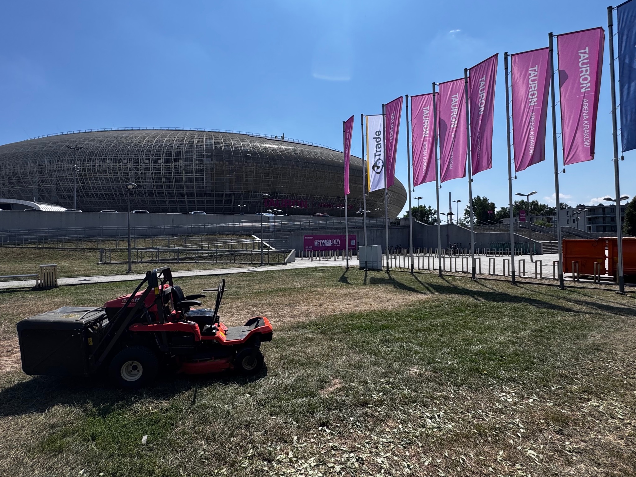 Czerwona kosiarka samojezdna z koszem na trawę na tle Tauron Areny Kraków i rzędu flag w słoneczny dzień. Częściowo skoszona trawa.
