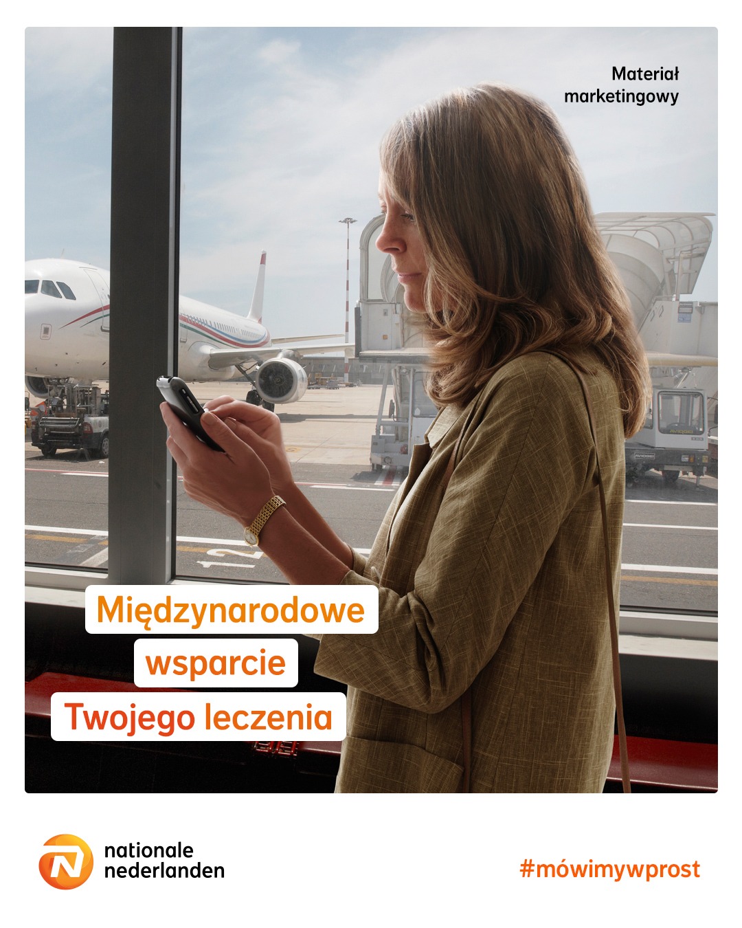 Kobieta w beżowej marynarce używa smartfona w hali lotniska, z widokiem na samolot przez okno.