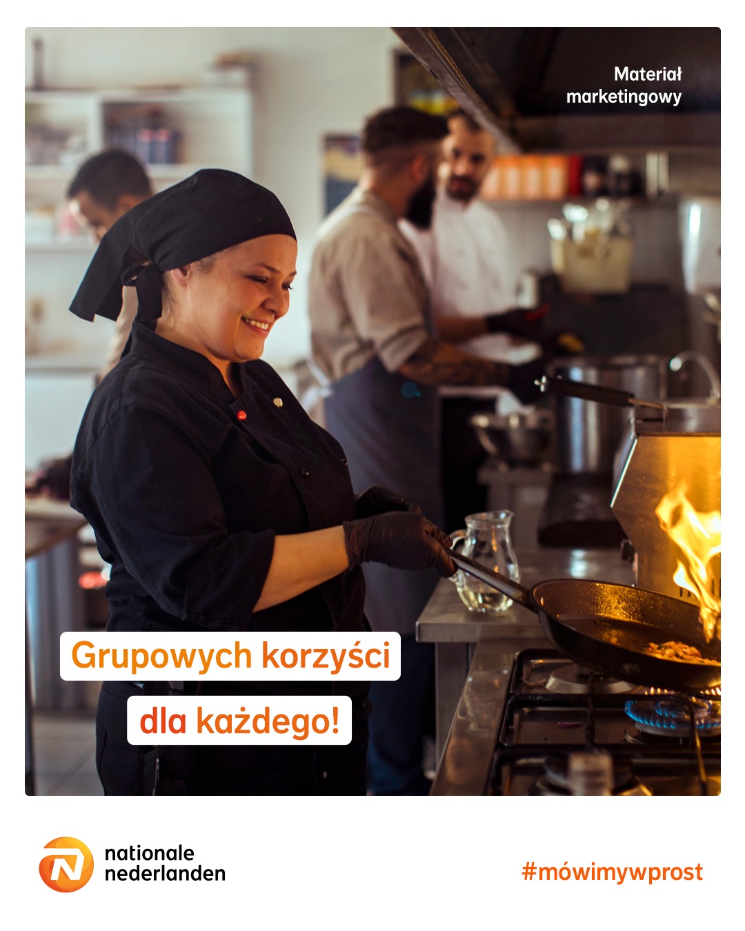 Uśmiechnięta kucharka w czarnym uniformie i rękawicach smaży potrawę na gazowym palniku w profesjonalnej kuchni; w tle inni kucharze i sprzęt kuchenny.