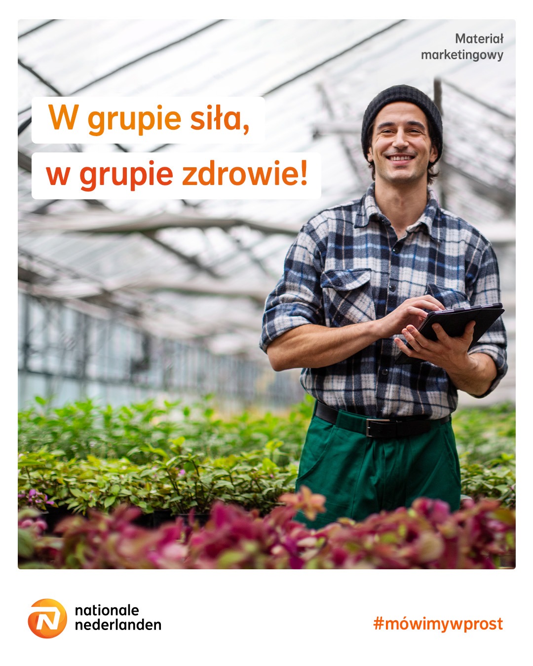 Uśmiechnięty mężczyzna w czapce i flanelowej koszuli, trzymający tablet w szklarni z roślinami, hasło 'W grupie siła, w grupie zdrowie!'