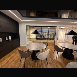 Baufirma Karpowicz - Nowoczesne wnętrze z drewnianą podłogą, okrągłymi stołami i krzesłami w stonowanych kolorach. Czarne lampy wiszące i przeszklenia dodają elegancji.