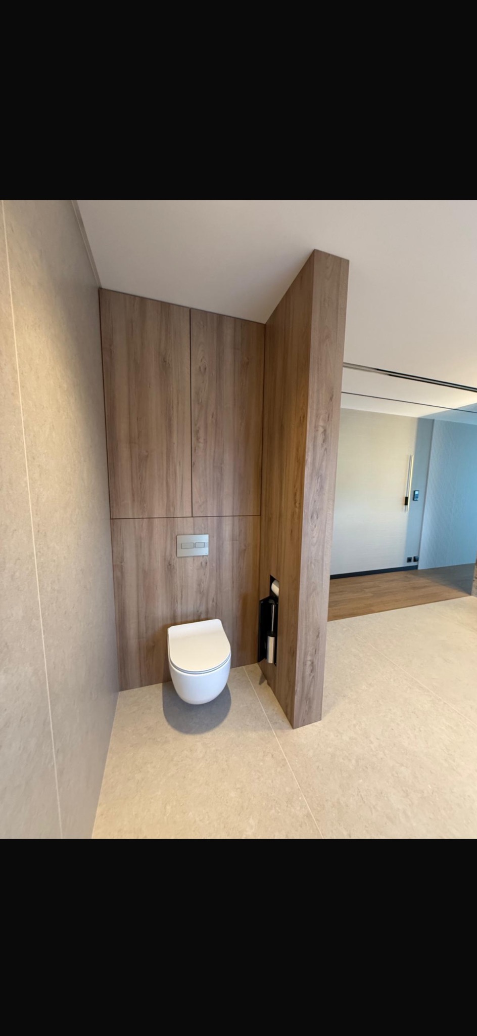 Nowoczesna toaleta podwieszana z zabudową z drewnopodobnej płyty, wkomponowana w minimalistyczną łazienkę z beżowymi płytkami i ukrytą rolką papieru toaletowego.