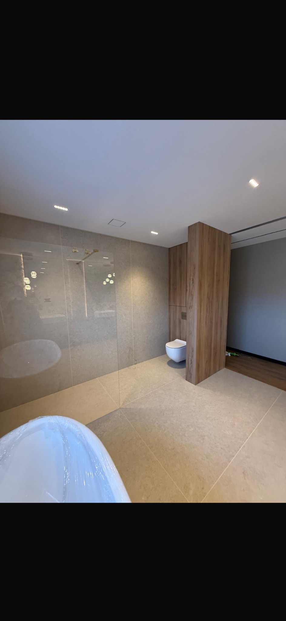 Nowoczesna łazienka z beżowymi płytkami, szklaną kabiną prysznicową i wolnostojącą wanną w folii ochronnej. Widoczna toaleta w zabudowie z drewnopodobnych paneli.