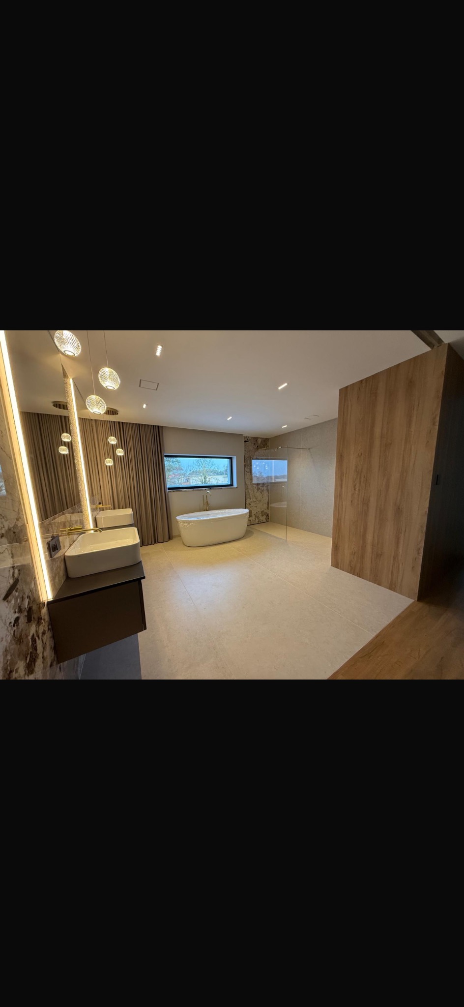 Przestronna łazienka z wolnostojącą wanną przy oknie, podwójną umywalką z lustrem i kabiną prysznicową. Beżowe płytki podłogowe i drewniane panele ścienne.