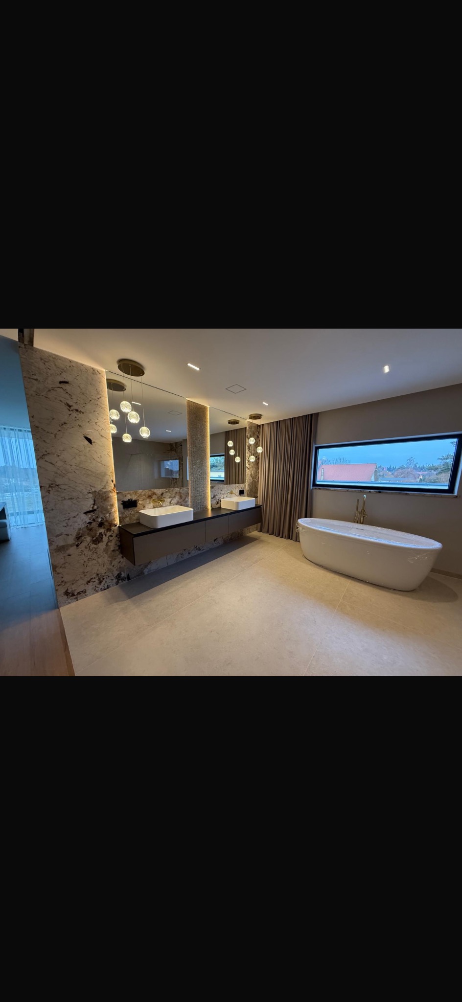 Nowoczesna łazienka z wanną wolnostojącą, dwoma umywalkami nablatowymi i dużym oknem z widokiem. Marmurowe wykończenie ściany i designerskie oświetlenie.