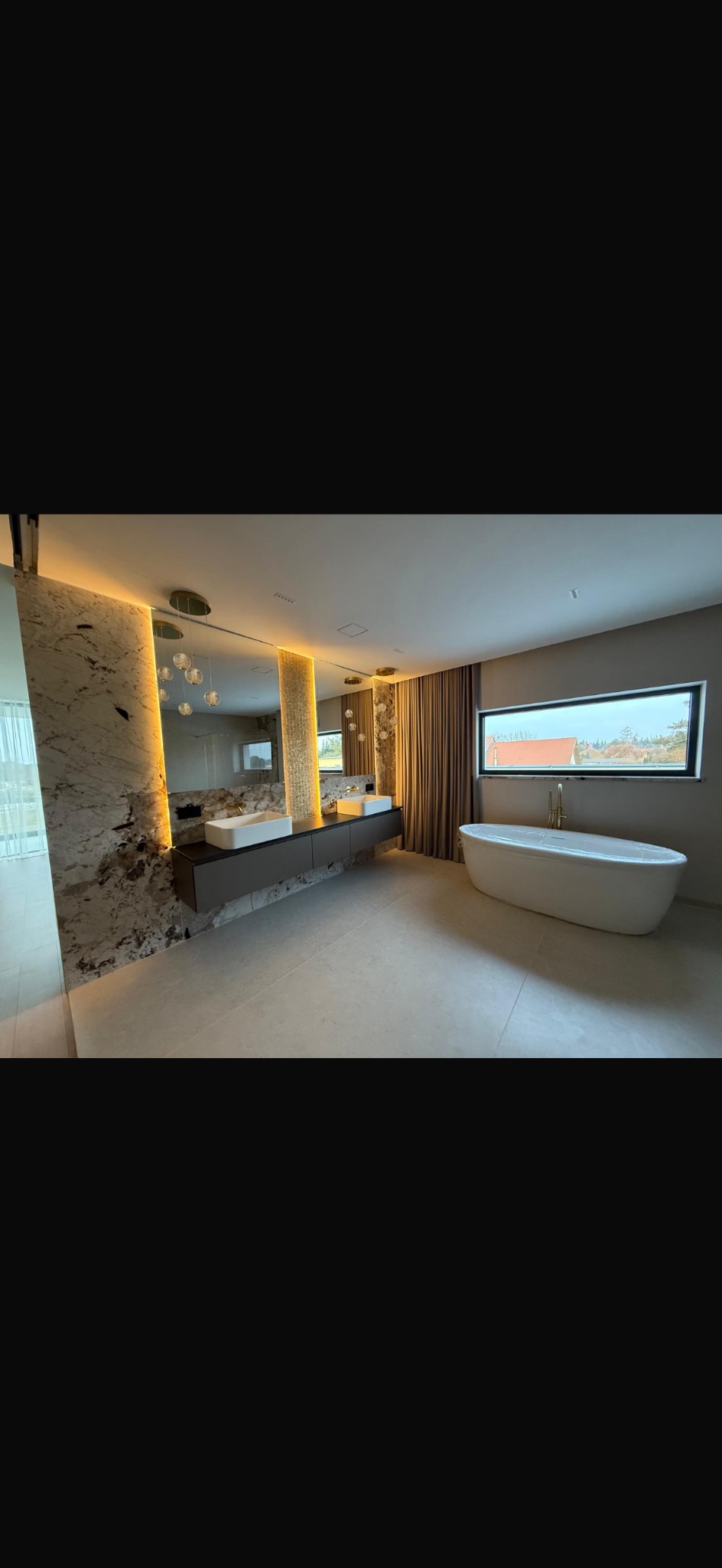 Nowoczesna łazienka z wanną wolnostojącą, podwójną umywalką na blacie i dużym lustrem z oświetleniem LED. Marmurowe wykończenie ściany i beżowa podłoga tworzą elegancki klimat.