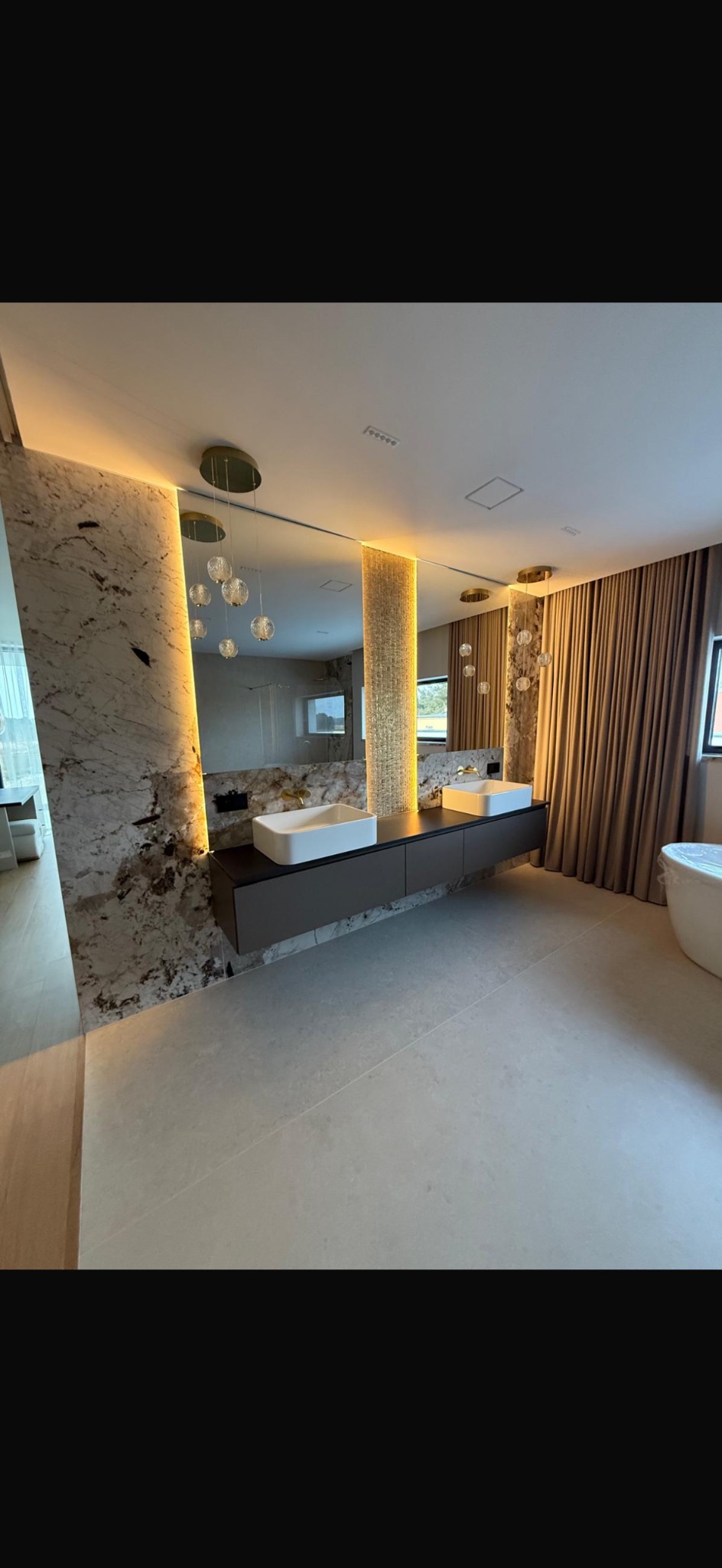 Nowoczesna łazienka z podwójną umywalką, marmurową ścianą z podświetleniem LED i dekoracyjnymi lampami wiszącymi. Minimalistyczny design z beżowymi zasłonami i wanną.