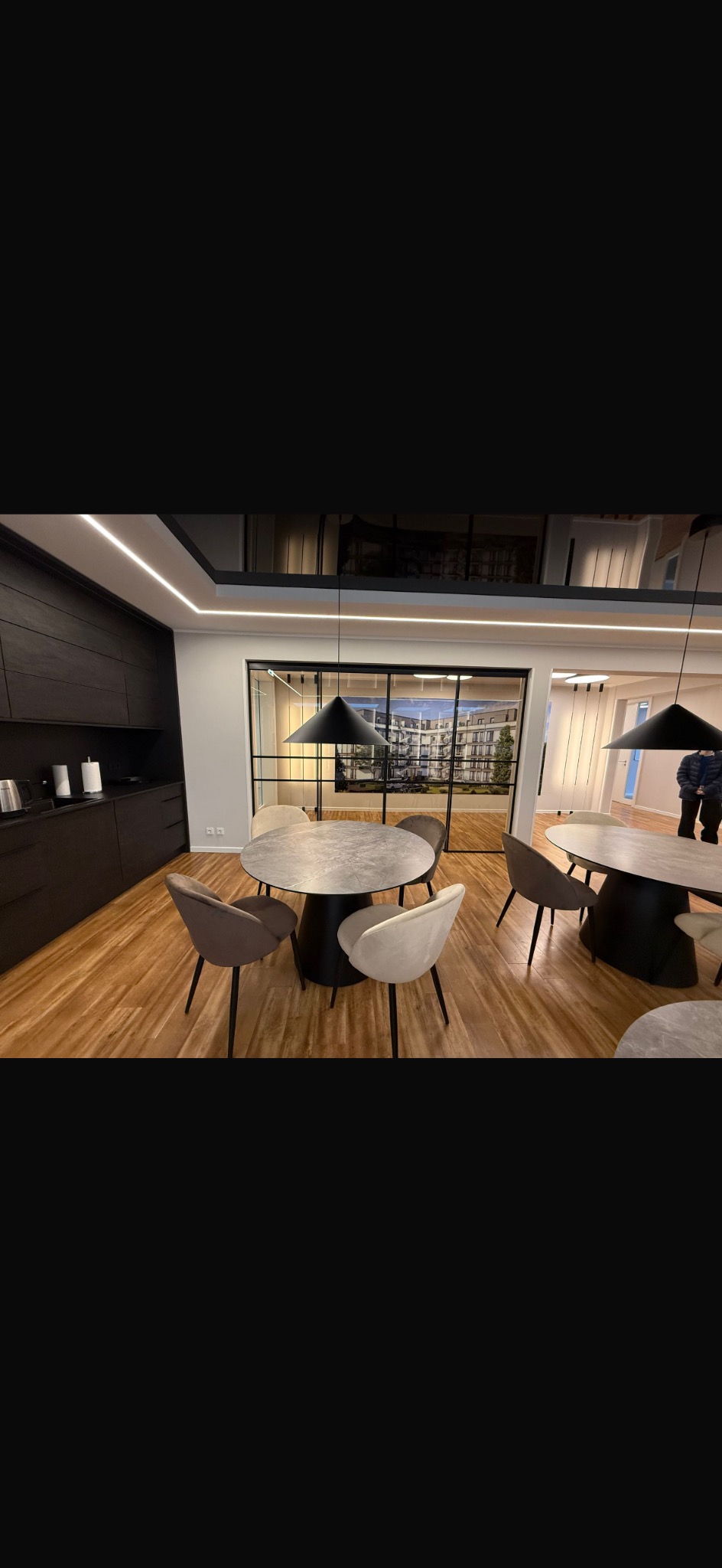 Nowoczesne wnętrze z drewnianą podłogą, okrągłymi stołami i krzesłami w stonowanych kolorach. Czarne lampy wiszące i przeszklenia dodają elegancji.