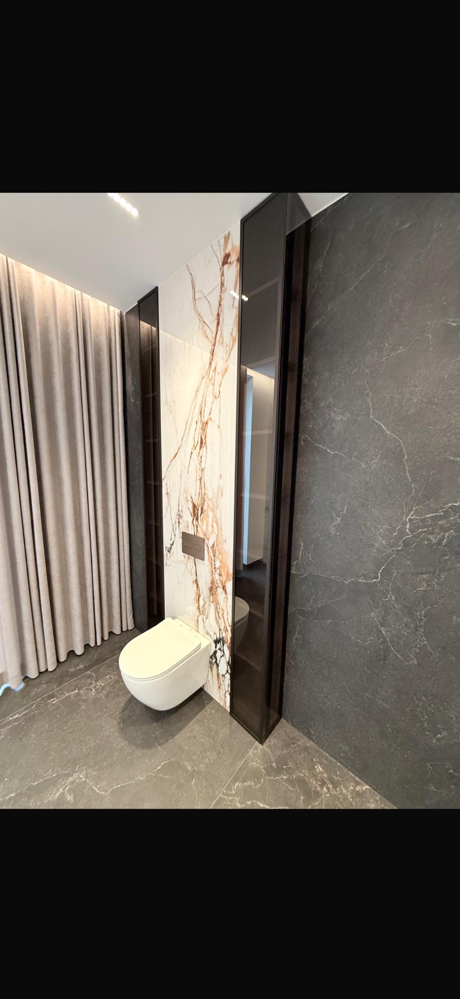 Nowoczesna toaleta podwieszana w łazience wykończonej marmurem i szarym gresem. Minimalistyczny design z zasłoną w tle i zabudową z ciemnego drewna.