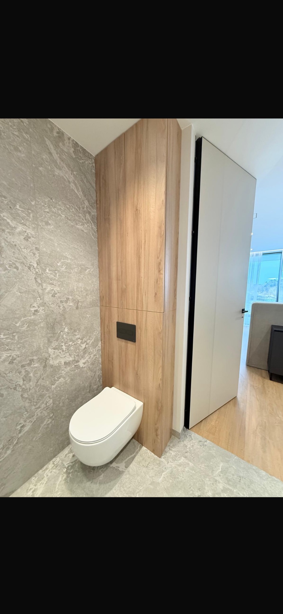 Nowoczesna toaleta podwieszana z zabudową z drewna, szara marmurowa ściana i podłoga, minimalistyczny design, widok na otwarte drzwi i dalszą część mieszkania.