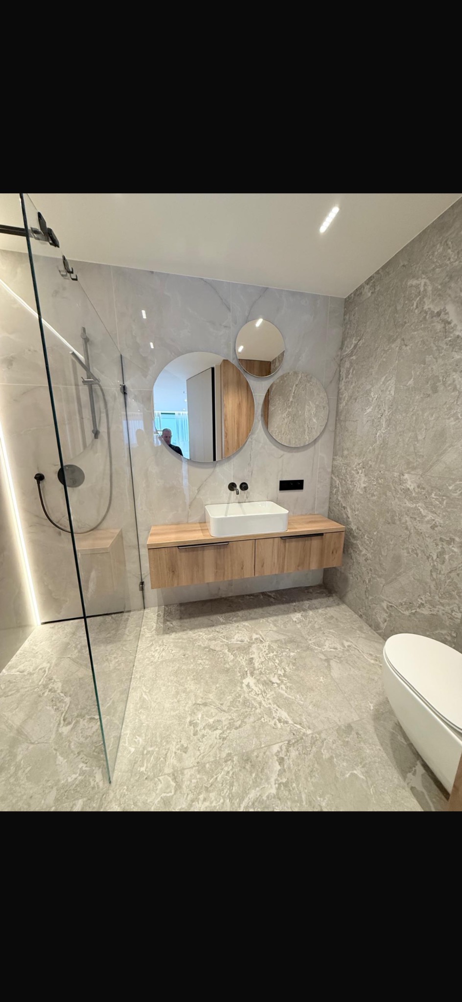 Nowoczesna łazienka z kabiną prysznicową typu walk-in, umywalką nablatową na drewnianej szafce i trzema okrągłymi lustrami na marmurowej ścianie.