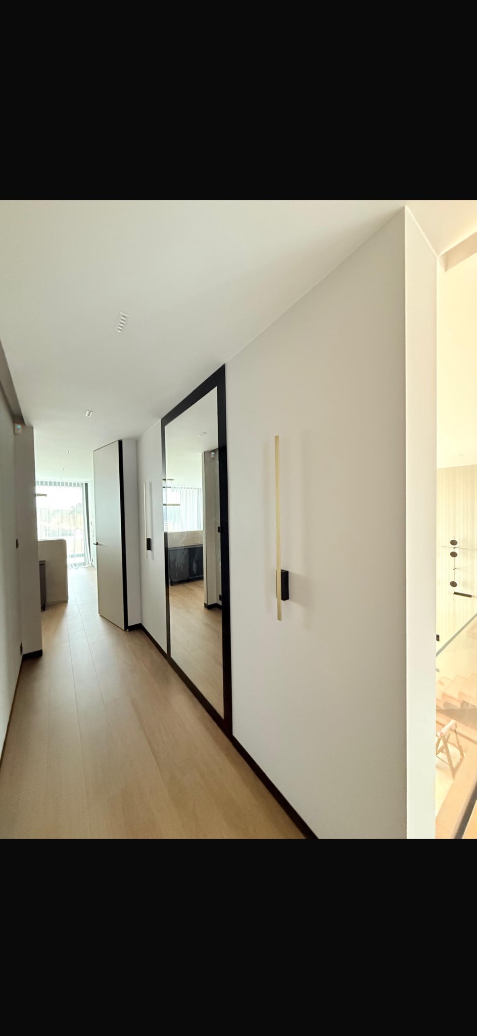Nowoczesny korytarz z lustrem w czarnej ramie i drewnianą podłogą. Minimalistyczne oświetlenie ścienne. Widok na salon z jasnymi meblami i balkon.