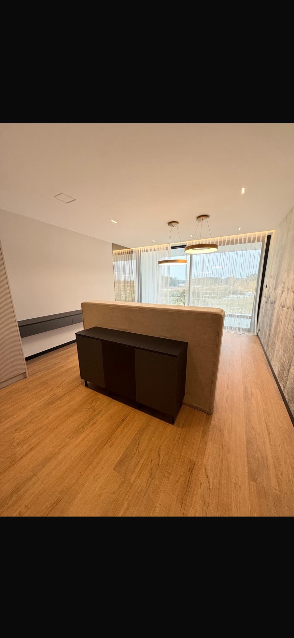 Nowoczesne wnętrze z drewnianą podłogą, szafką RTV i beżową ścianką działową. Widoczne okno z zasłonami i designerskie lampy sufitowe.