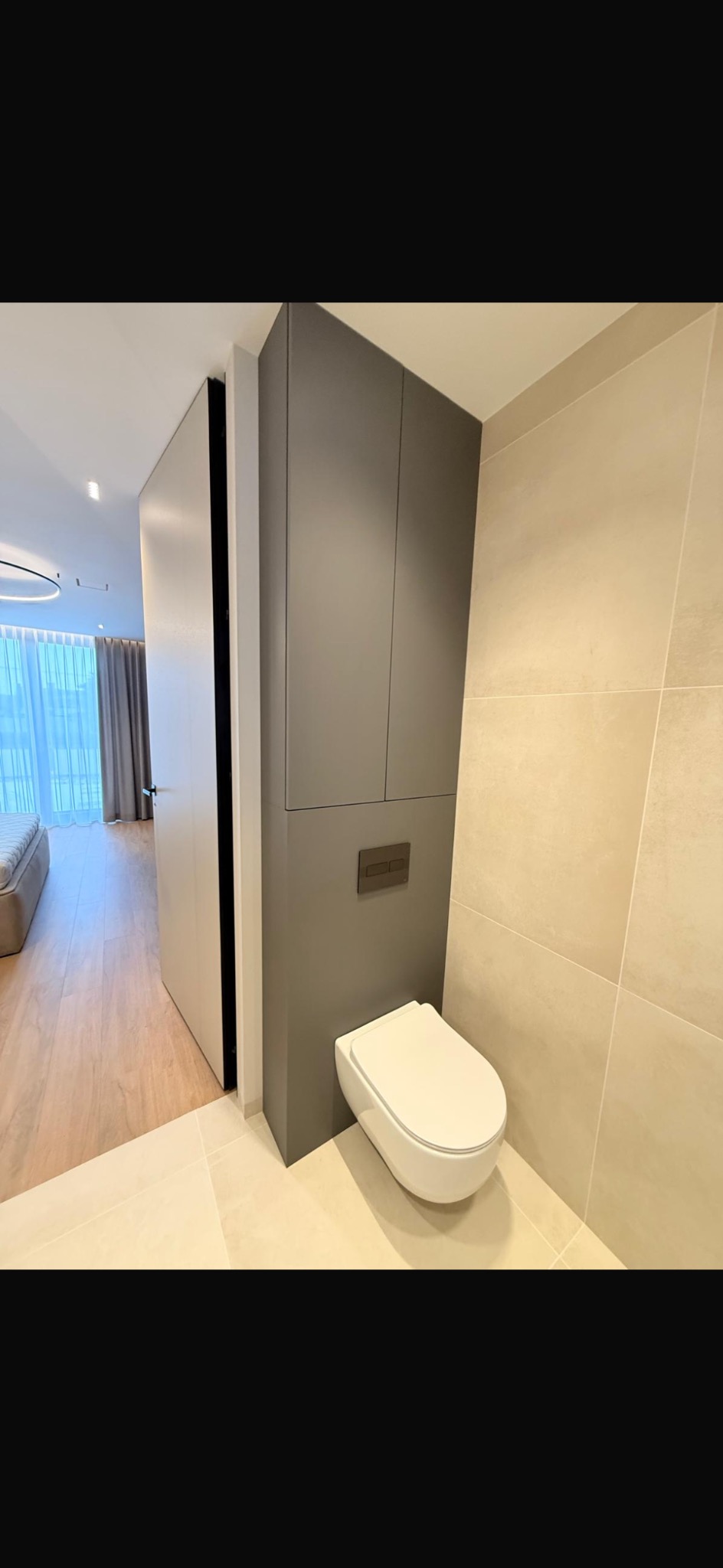 Nowoczesna łazienka z podwieszaną toaletą i szafką w zabudowie, beżowe płytki, widok na sypialnię przez otwarte drzwi. Minimalistyczny design, neutralna kolorystyka.