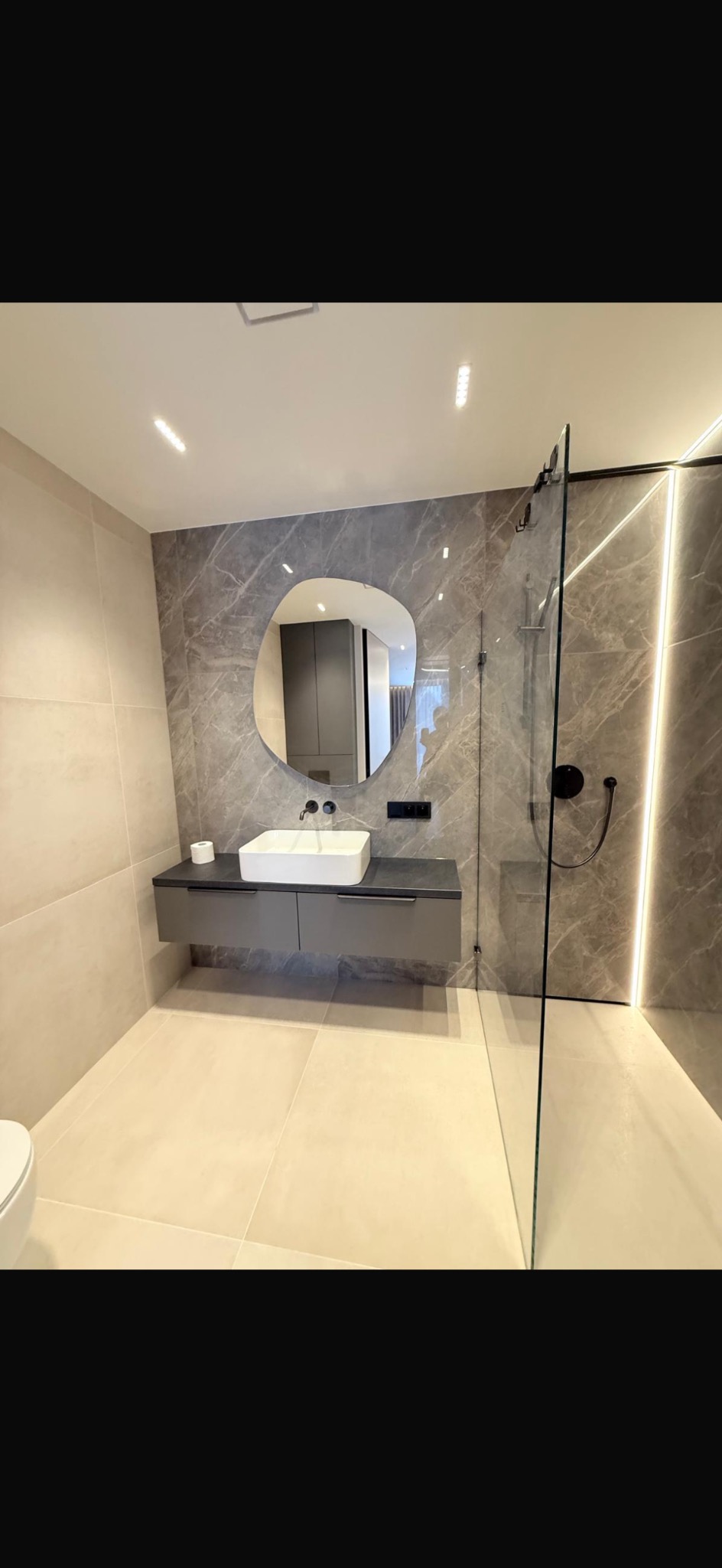 Nowoczesna łazienka z marmurowymi płytkami, asymetrycznym lustrem i kabiną prysznicową typu walk-in. Minimalistyczny design, beżowe płytki podłogowe i subtelne oświetlenie LED.