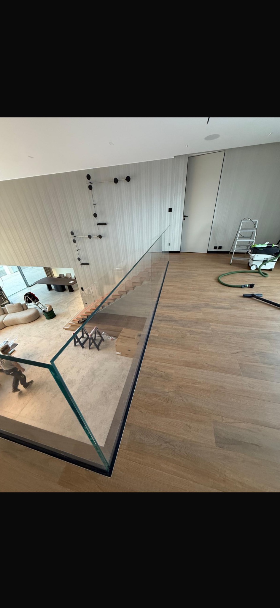 Nowoczesne wnętrze z drewnianą podłogą i szklaną balustradą schodów. Widoczny odkurzacz i drabina, sugerujące prace wykończeniowe. Minimalistyczny design.