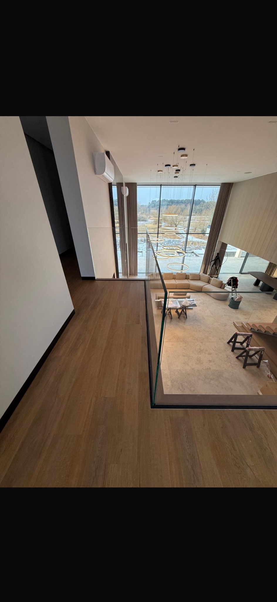 Nowoczesne wnętrze domu z dużym oknem i widokiem na zewnątrz. Jasne ściany, drewniana podłoga i szklana balustrada. W tle sofa i elementy dekoracyjne.