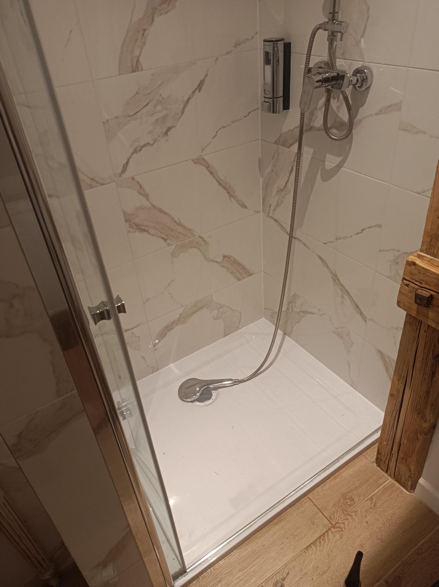 Nowoczesny prysznic z białą brodzikiem, chromowaną armaturą i marmurkowymi płytkami ściennymi, uzupełniony o element z surowego drewna.