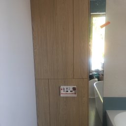 SoBo Meble wytwórnia meble indywidualnych - Drewniana zabudowa stelaża WC z przyciskiem spłukującym i elementami instalacji, widoczna wanna w tle.