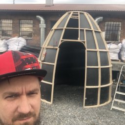 SoBo Meble wytwórnia meble indywidualnych - Mężczyzna w czapce robi selfie na tle szkieletu konstrukcji w kształcie igloo z drewnianych belek i czarnych paneli, w tle budynek z cegły i worki z materiałami budowlanymi.