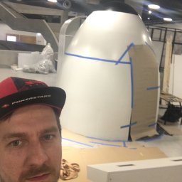 SoBo Meble wytwórnia meble indywidualnych - Selfie mężczyzny w czapce PokerStars na tle dużej, białej, jajowatej konstrukcji z niebieską taśmą malarską i kartonem, prawdopodobnie w trakcie budowy w przestronnym, industrialnym wnętrzu.
