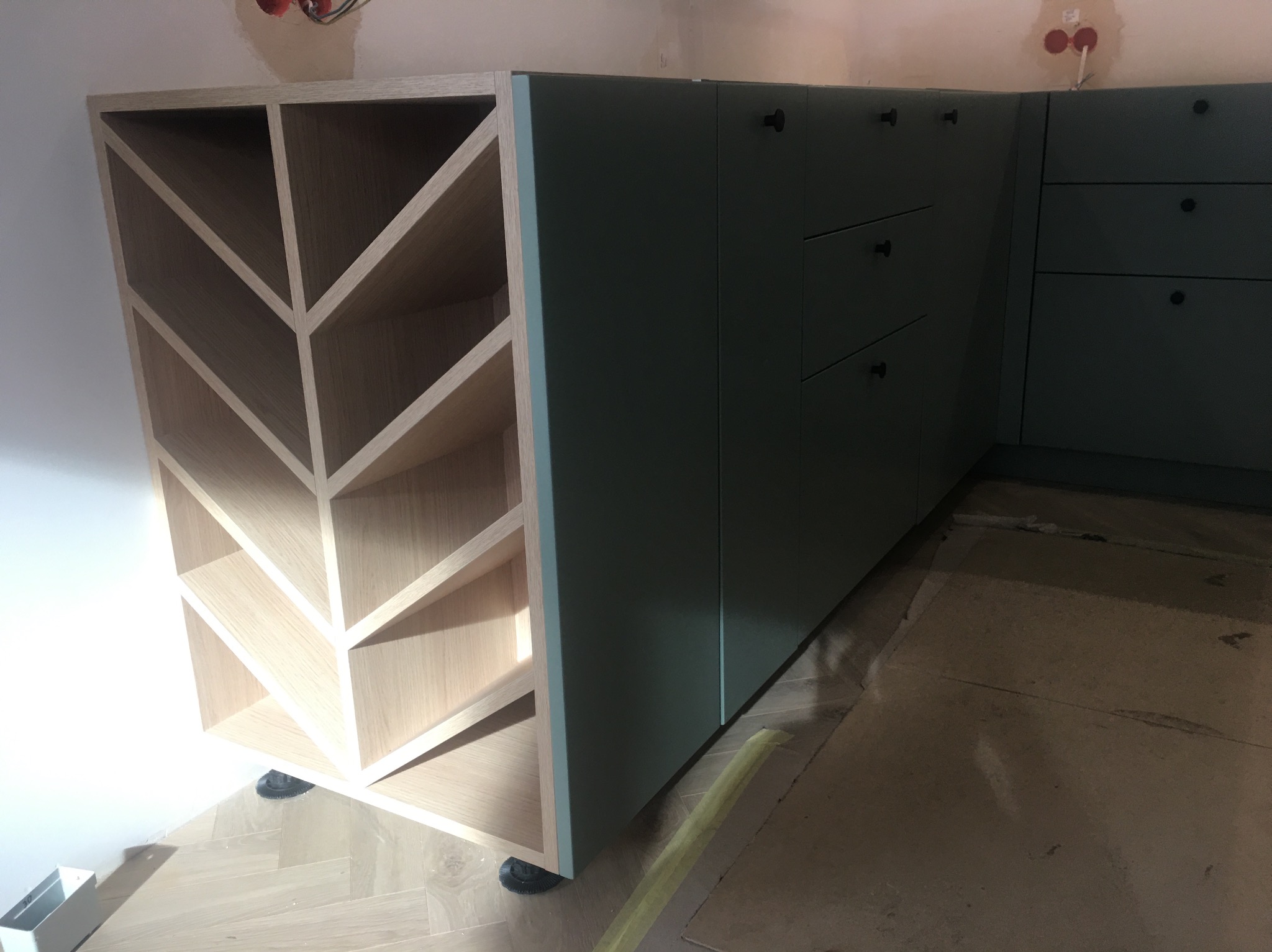 Fragment montażu kuchni na wymiar: szafka z drewnianymi przegródkami o wzorze strzałek i zielone szafki z czarnymi gałkami na tle surowej podłogi.