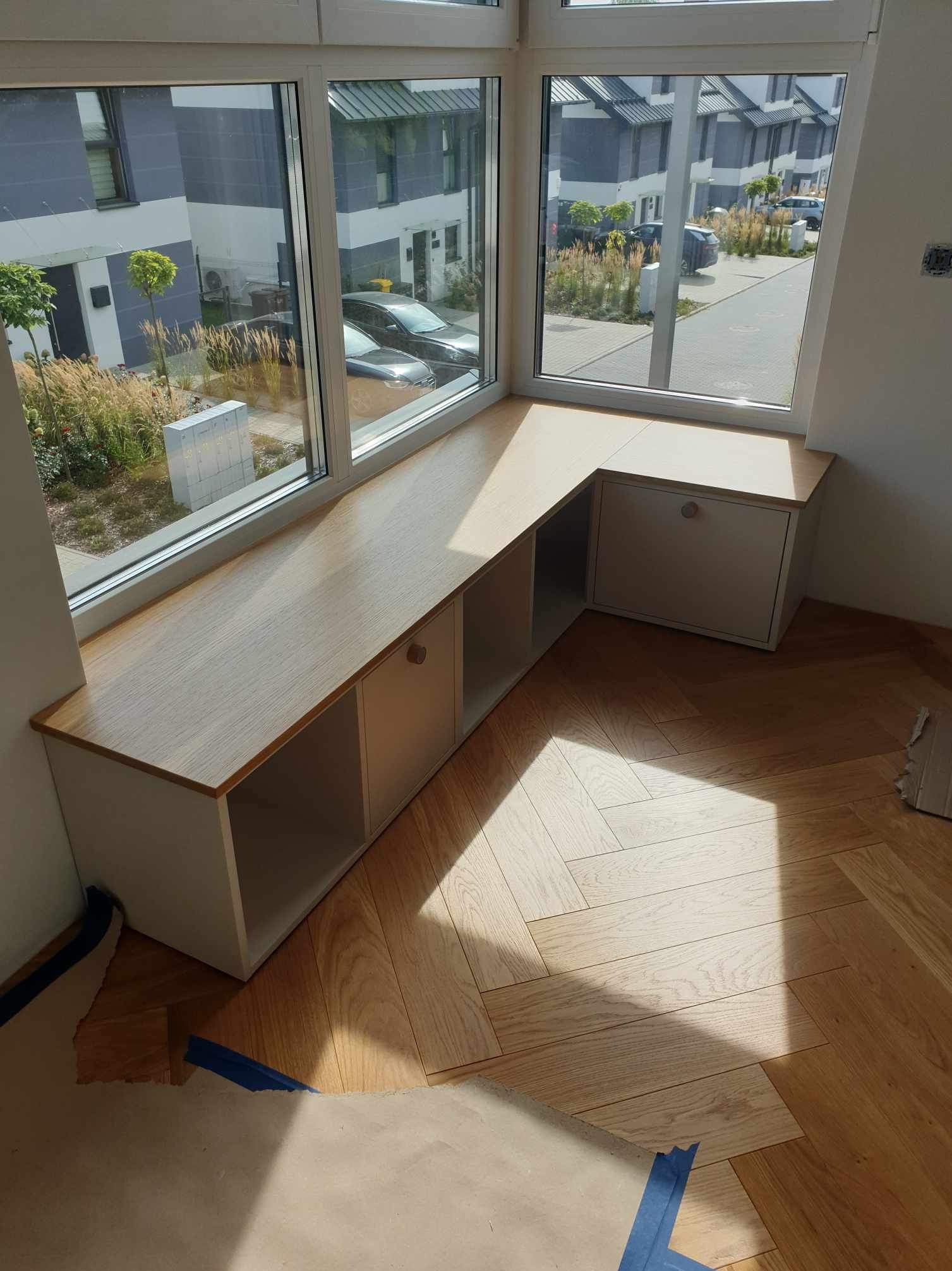 Zabudowa okienna z drewnianym blatem i szafkami, widok z góry na parkiet w jodełkę, za oknem widok na osiedle domów.