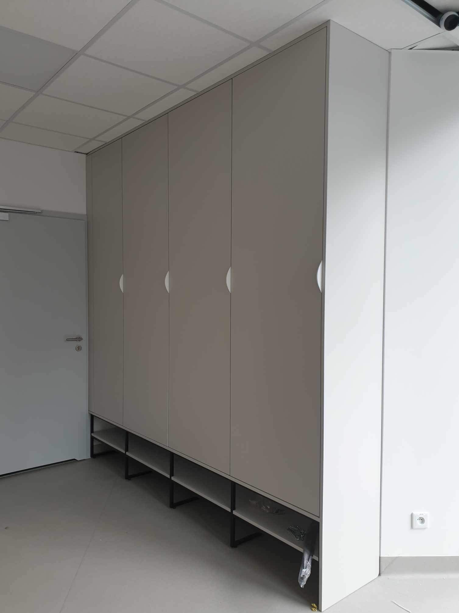 Duża, szara szafa wnękowa z półkami na metalowym stelażu, ustawiona w pomieszczeniu z białą ścianą i szarą podłogą, widoczne elementy montażowe na podłodze.